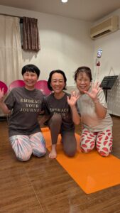 オレンジ色のヨガマットの上で「EMBRACE YOUR JOURNEY」のお揃いTシャツを着て笑顔で手を振る3人の女性